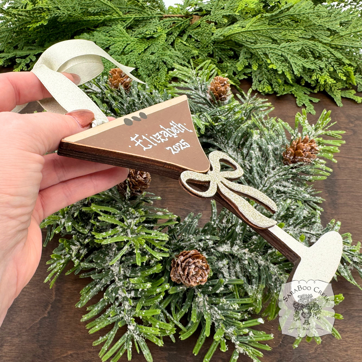 Hand holding a wooden espresso martini ornament with 'Elizabeth 2025' handwritten in white with a glittered silver wood bow; hung with cream satin ribbon