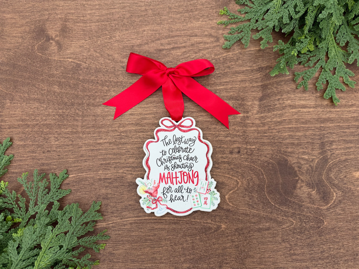 A Christmas ornament shaped like a gift box, decorated with a red ribbon bow and printed with the text 'The brightest way to celebrate Christmas cheer is sharing MAHjong for all to see!' in black and red lettering, displayed on a wooden surface with green pine branches.