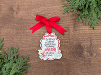 A Christmas ornament shaped like a gift box, decorated with a red ribbon bow and printed with the text 'The brightest way to celebrate Christmas cheer is sharing MAHjong for all to see!' in black and red lettering, displayed on a wooden surface with green pine branches.
