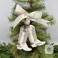 Decorative wood Christmas ornament with white cowboy boots hung by a cream satin bow on a tree.